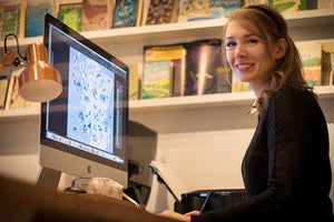 Sara Mulvanny sitting in front of an imac with books on a shelf in the background