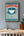 Framed Alice In Wonderland poster featuring a  teacup design on a wall above a wooden desk with a lamp and books.