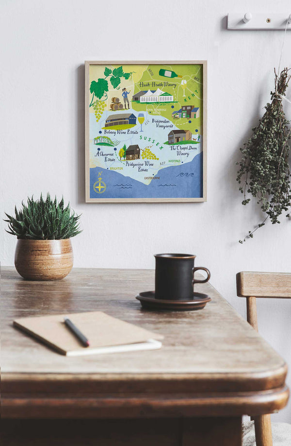 Framed map of English wine estates in Sussex and Kent on a wall above a wooden table with a mug, notebook, and pen.