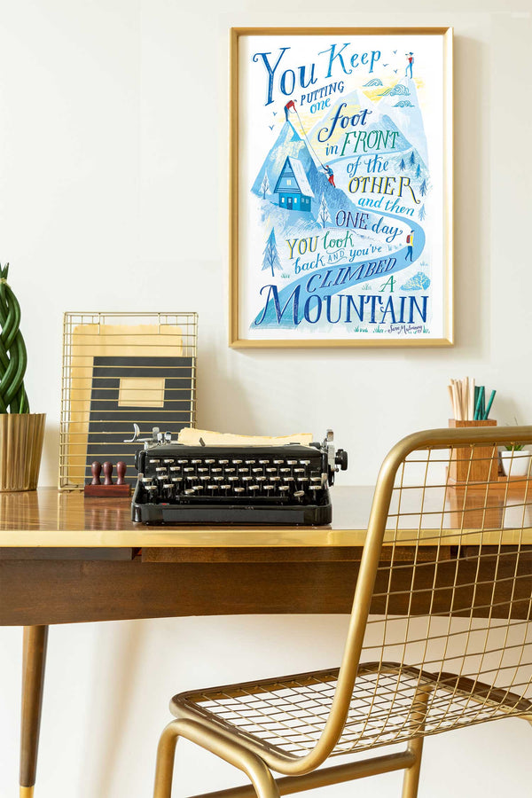 Framed artwork with motivational quote above a desk with a typewriter and chair.