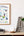 Framed office ergonomics poster on a wall above a wooden shelf with books, a plant, and decorative letters.