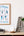 Framed Office Stretches poster on a wall above a wooden shelf with books, a plant, and decorative letters.