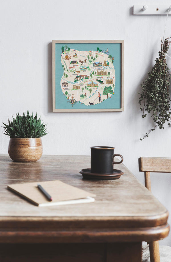 Framed map of Stamford region on a wall above a wooden table with a cup, notebook, and pen. Illustrated map features Rutland, Market Harborough, Uppingham, Oundle, Nene Valley, Melton Mowbray and Oakham
